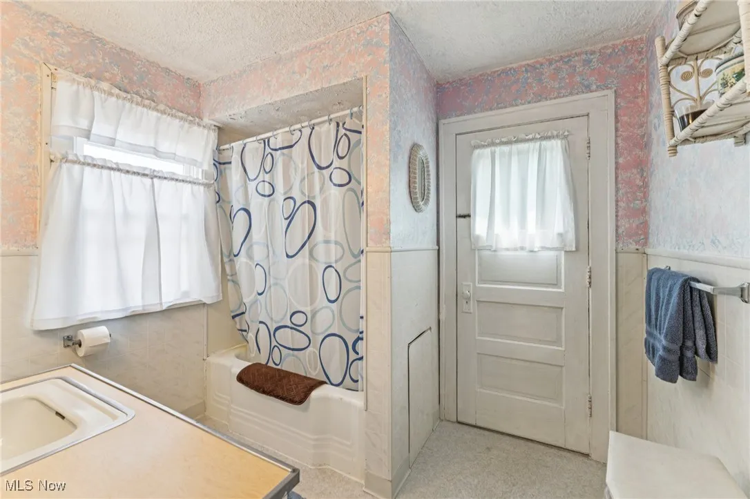 Full bathroom with shower / bathtub combination with curtain, tile walls, a textured ceiling, wainscoting, and wallpapered walls
