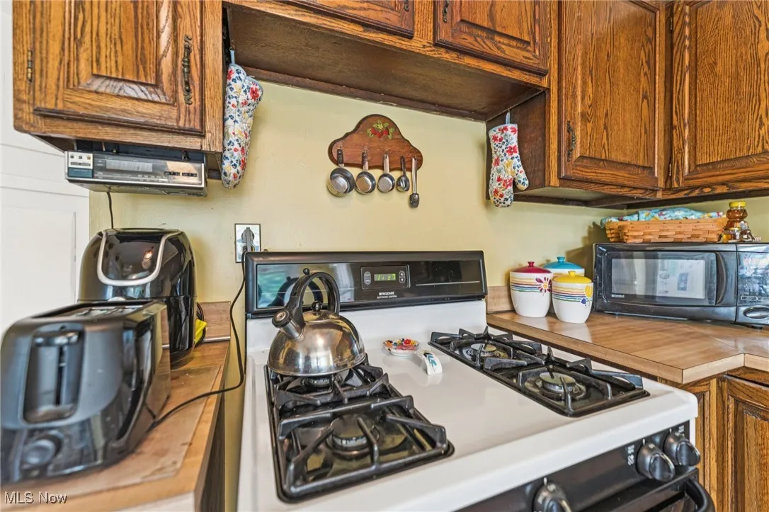 Kitchen with white gas stove, black microwave, light countertops, brown cabinetry, and under cabinet range hood