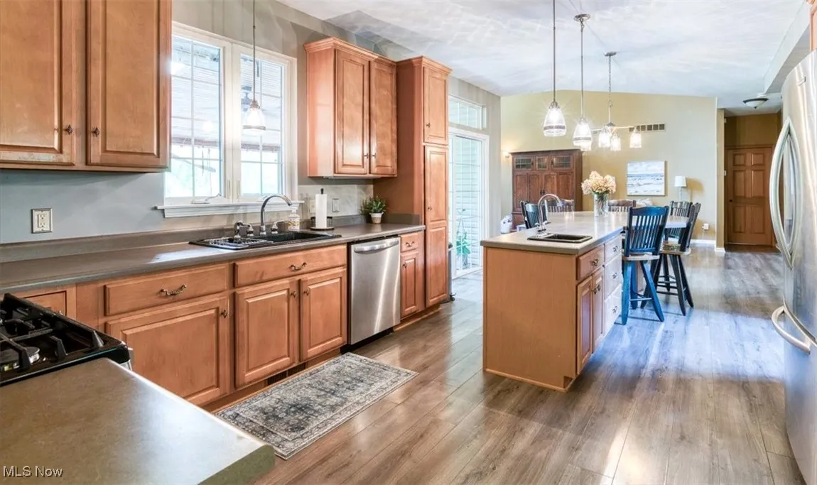 Kitchen featuring an island with sink, brown cabinets, dark wood-style flooring, hanging light fixtures, and stainless steel appliances