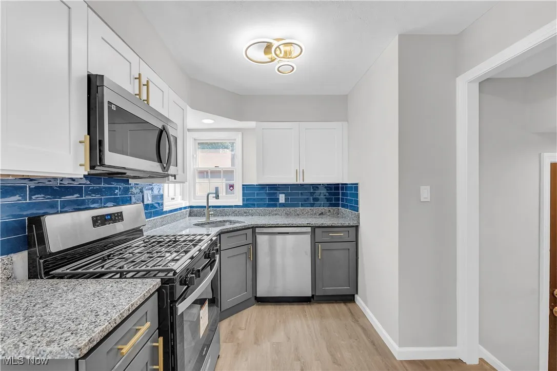 Kitchen with stainless steel appliances, gray cabinets, light stone counters, backsplash, and white cabinets