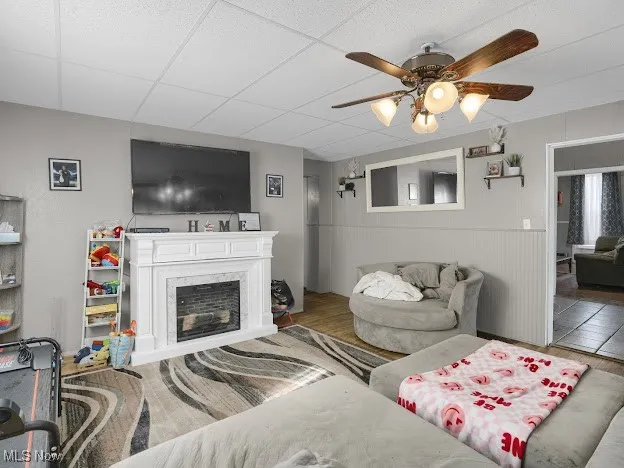 Living room with ceiling fan, wood finished floors, a drop ceiling, a high end fireplace, and a wainscoted wall