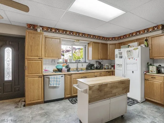 Kitchen with white refrigerator with ice dispenser, a paneled ceiling, stainless steel dishwasher, decorative backsplash, and a kitchen island