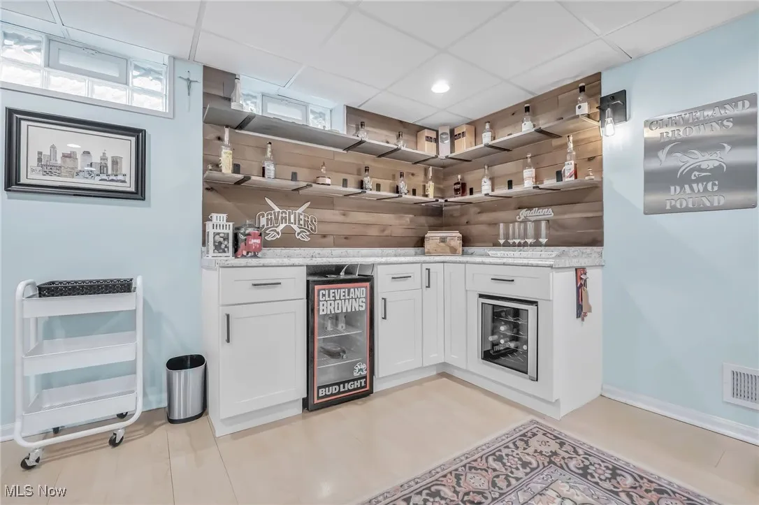 Bar with white cabinets, a paneled ceiling, wine cooler, open shelves, and light stone countertops
