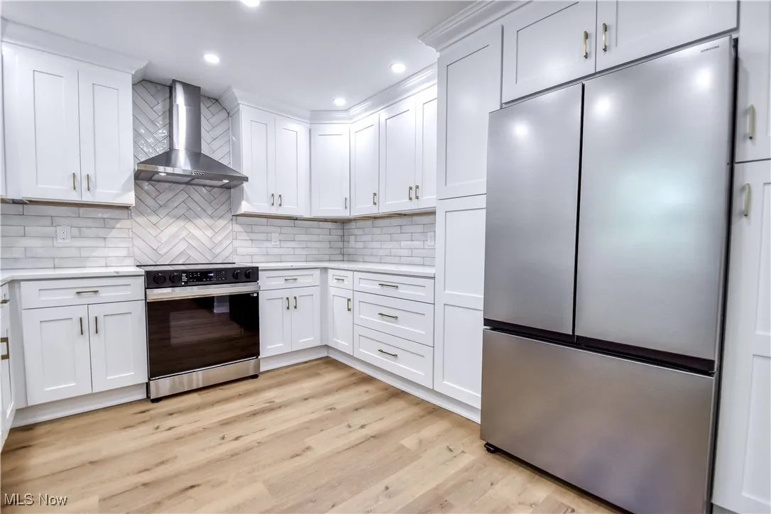Kitchen with freestanding refrigerator, range with electric cooktop, wall chimney range hood, white cabinetry, and light wood-style floors