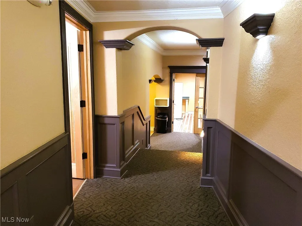Corridor with wainscoting, a decorative wall, crown molding, carpet floors, and arched walkways