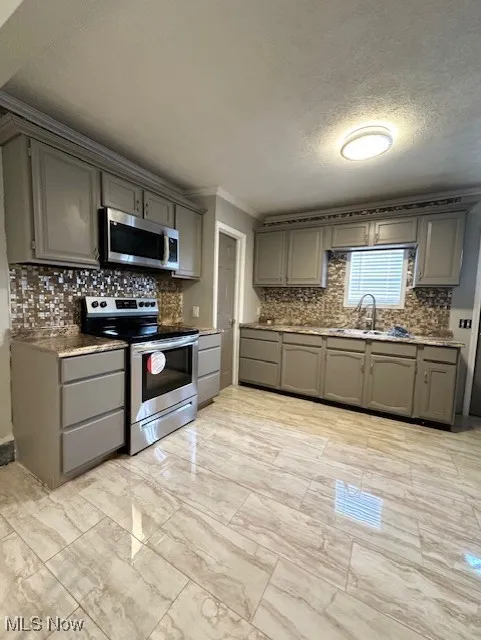 Kitchen featuring gray cabinetry, stainless steel appliances, decorative backsplash, crown molding, and a textured ceiling