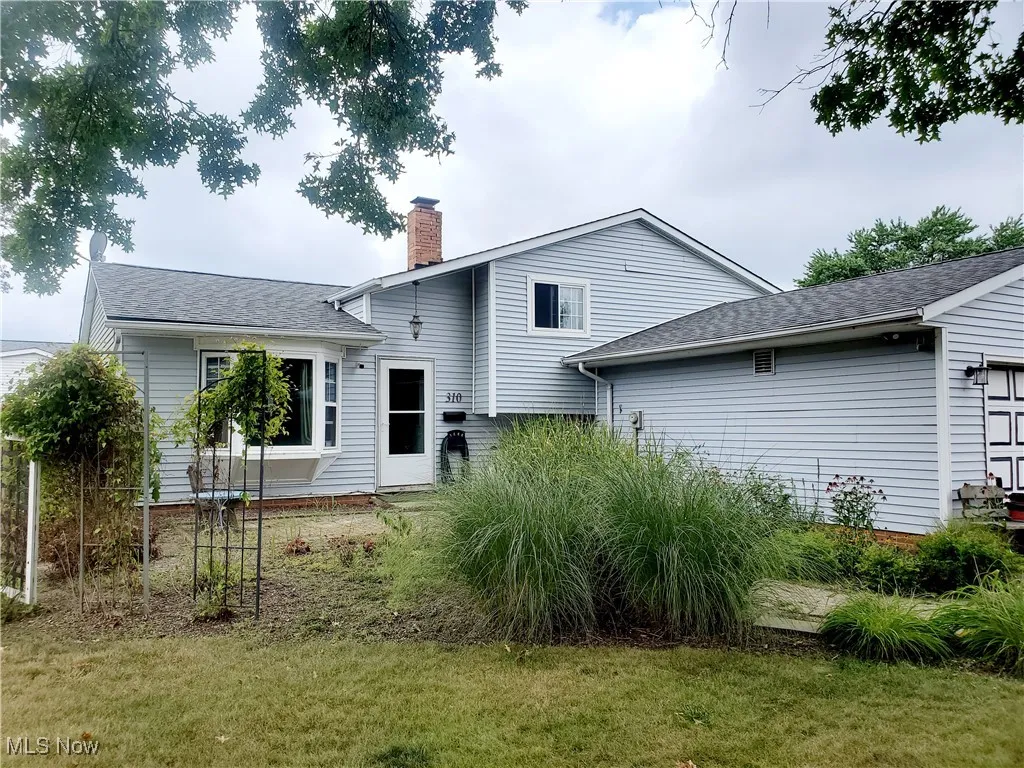 Rear view of house with a garage, a shingled roof, a chimney, and a lawn