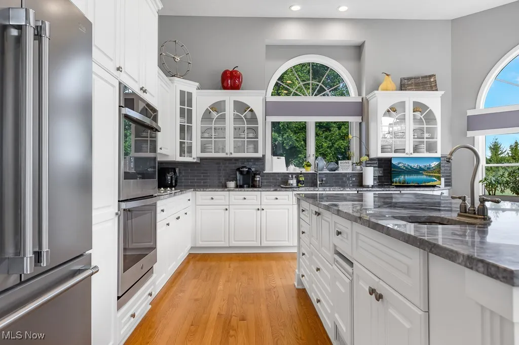 Kitchen with appliances with stainless steel finishes, glass insert cabinets, light wood-type flooring, tasteful backsplash, and dark stone counters