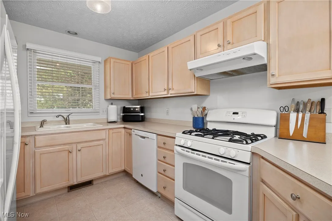 Kitchen featuring light brown cabinets, white appliances, under cabinet range hood, light countertops, and a textured ceiling
