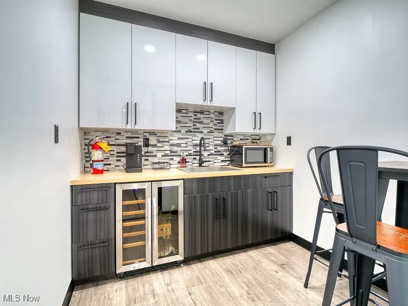 Indoor wet bar with white cabinetry, beverage cooler, backsplash, light wood-type flooring, and modern cabinets