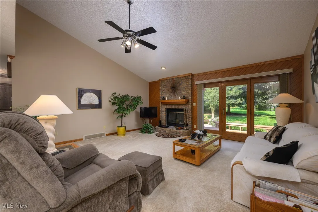 Carpeted living room with lofted ceiling, a textured ceiling, a brick fireplace, ceiling fan, and wooden walls