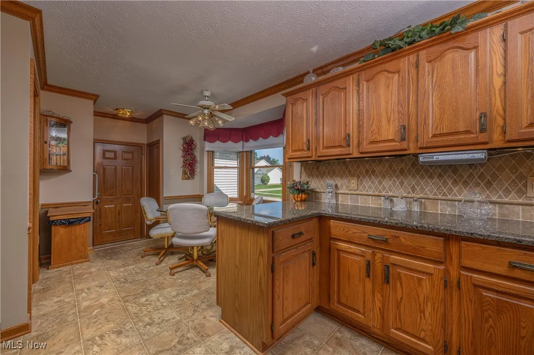 Kitchen featuring brown cabinets, backsplash, dark stone countertops, ceiling fan, and a wainscoted wall