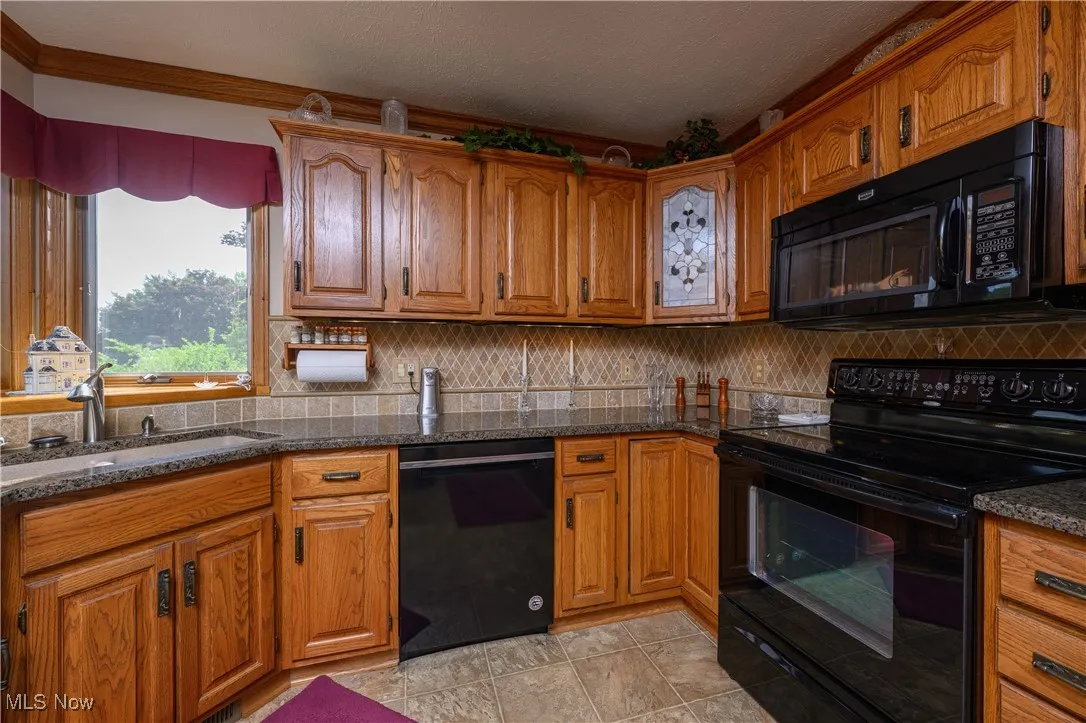 Kitchen with black appliances, brown cabinetry, backsplash, dark stone counters, and ornamental molding