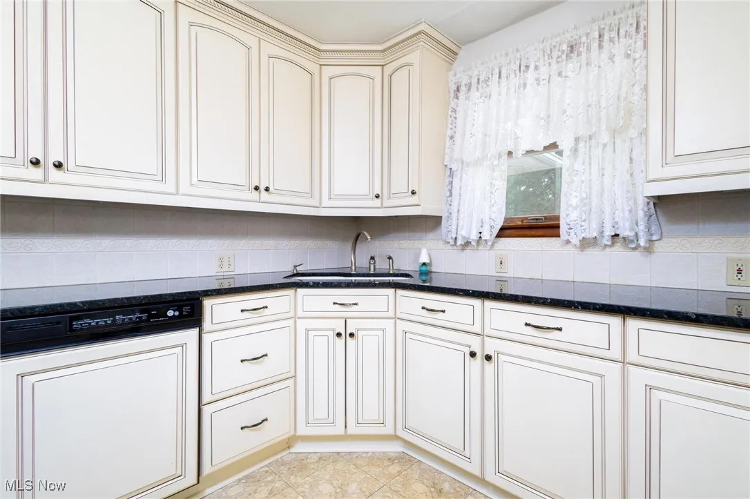 Kitchen with dishwasher, tasteful backsplash, dark stone countertops, cream cabinetry, and light tile patterned flooring