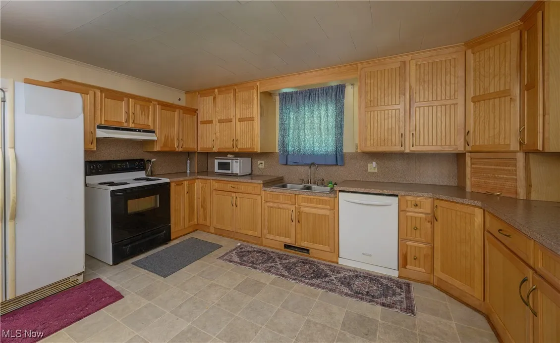Kitchen featuring white appliances, decorative backsplash, under cabinet range hood, light brown cabinetry, and crown molding