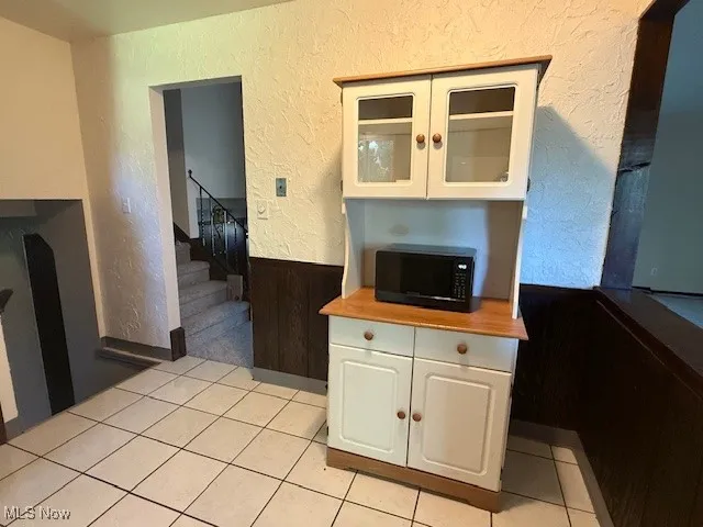 Kitchen with a textured wall, white cabinetry, glass insert cabinets, light tile patterned floors, and a wainscoted wall