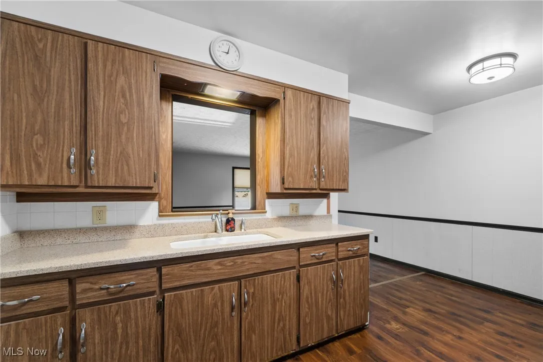 Kitchen with dark wood-style flooring, wainscoting, light stone counters, decorative backsplash, and brown cabinetry