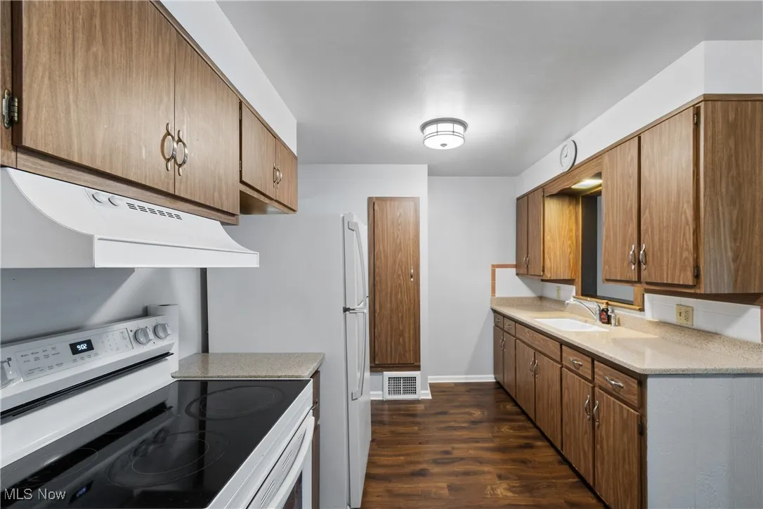 Kitchen with white appliances, under cabinet range hood, dark wood-style flooring, light stone counters, and brown cabinets