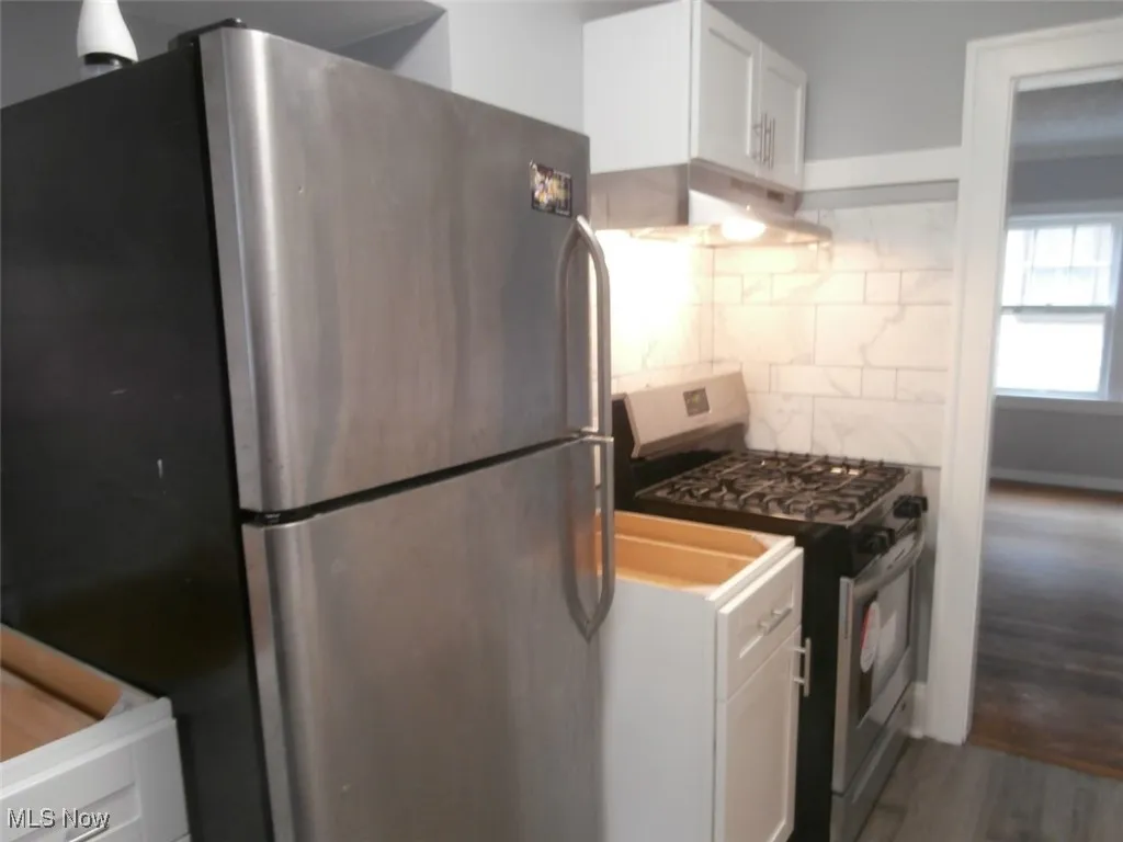 Kitchen with white cabinets, stainless steel appliances, decorative backsplash, under cabinet range hood, and dark wood-style flooring