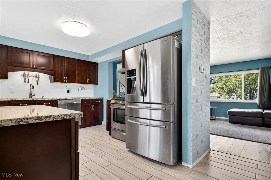 Kitchen with dark brown cabinetry, stainless steel appliances, light wood-style floors, light stone countertops, and a textured ceiling