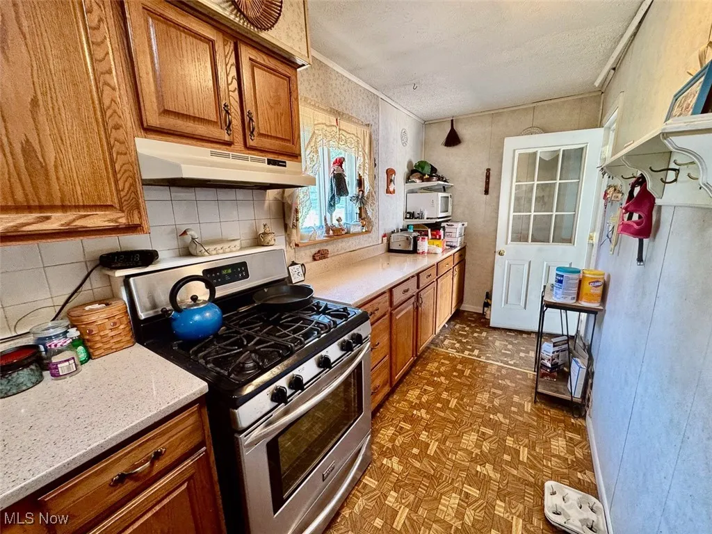 Kitchen with gas range, brown cabinetry, a textured ceiling, under cabinet range hood, and tasteful backsplash