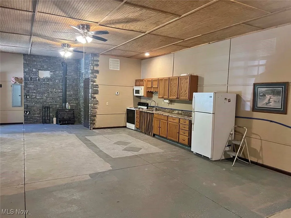 Kitchen featuring a wood stove, white appliances, ceiling fan, brown cabinets, and concrete flooring