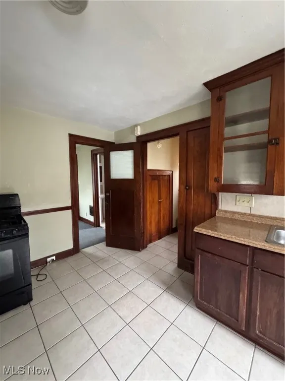 Kitchen featuring stove, light tile patterned floors, dark brown cabinets, light stone counters, and glass insert cabinets
