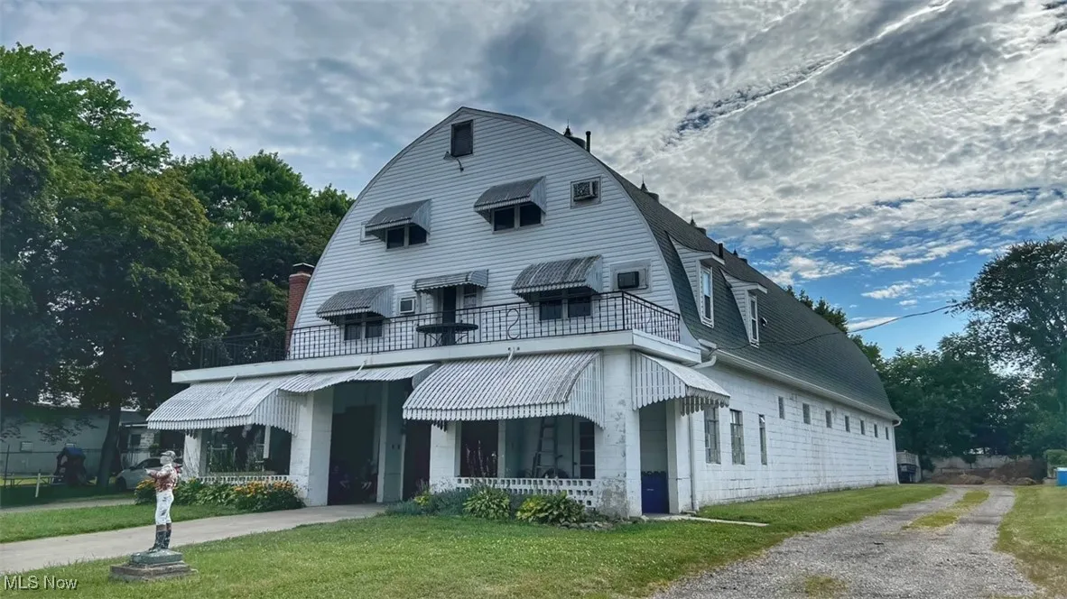 View of front facade featuring gravel driveway, a gambrel roof, a porch, a front lawn, and a chimney