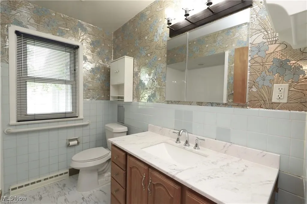 Bathroom with wallpapered walls, a baseboard heating unit, vanity, tile walls, and a wainscoted wall