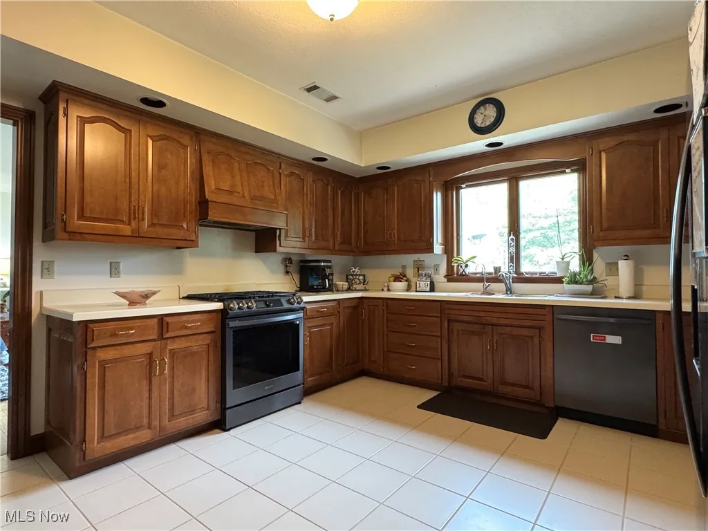 Kitchen featuring light countertops, stainless steel appliances, light tile patterned floors, brown cabinetry, and custom range hood