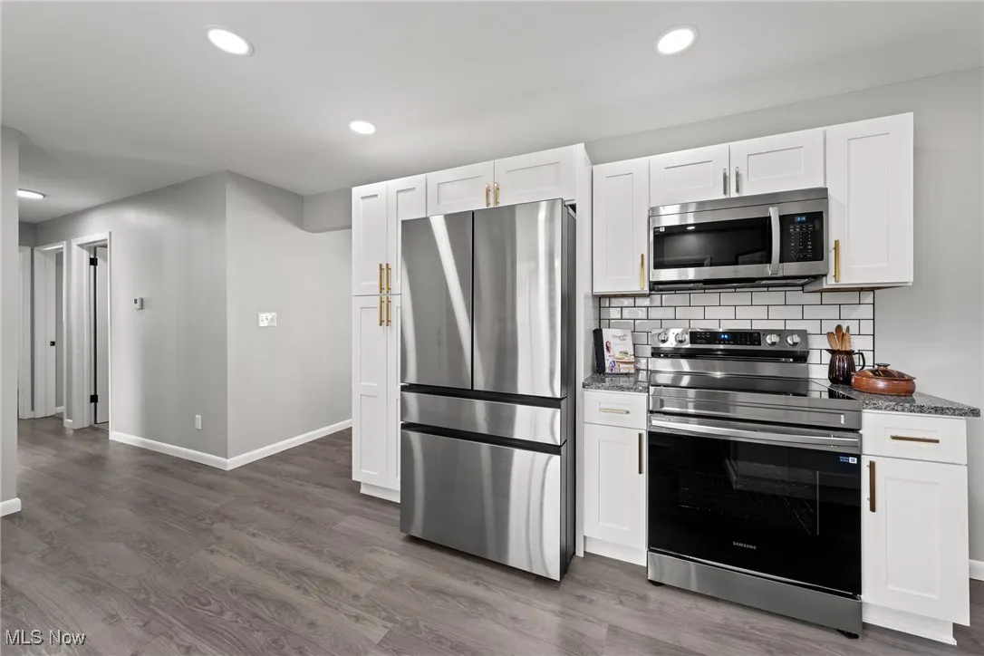 Kitchen with appliances with stainless steel finishes, tasteful backsplash, dark stone countertops, white cabinets, and dark wood-style flooring