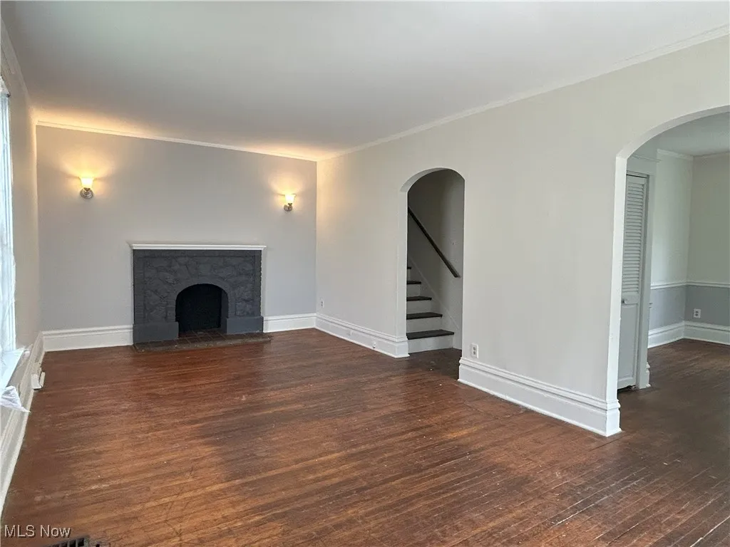 Unfurnished living room with a fireplace with raised hearth, dark wood finished floors, arched walkways, stairs, and ornamental molding