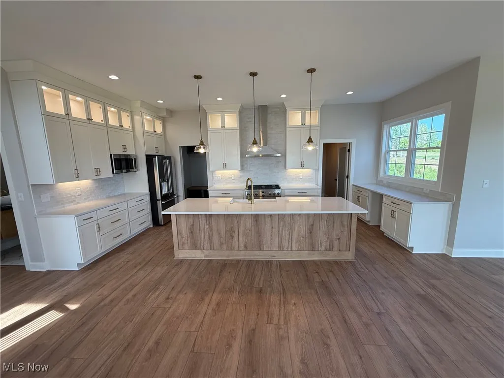 Kitchen with white cabinetry, an island with sink, decorative light fixtures, wall chimney range hood, and glass insert cabinets