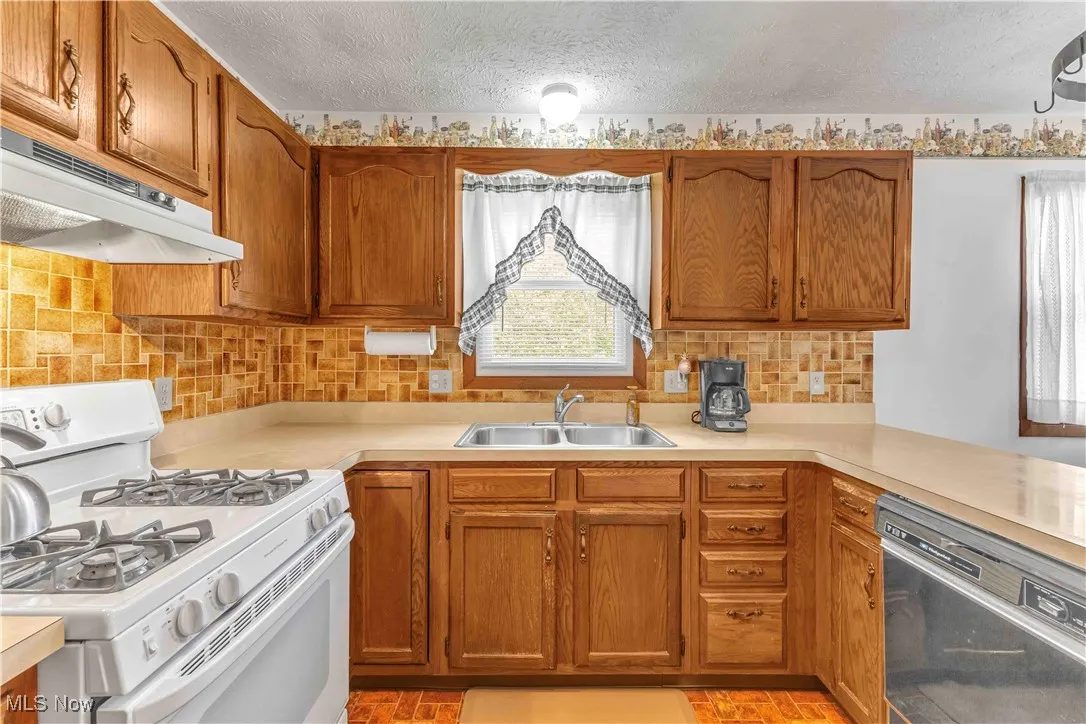 Kitchen with solid, wood cabinetry, white gas stove, white overhead range fan, kitchen sink with window above and light colored counters.