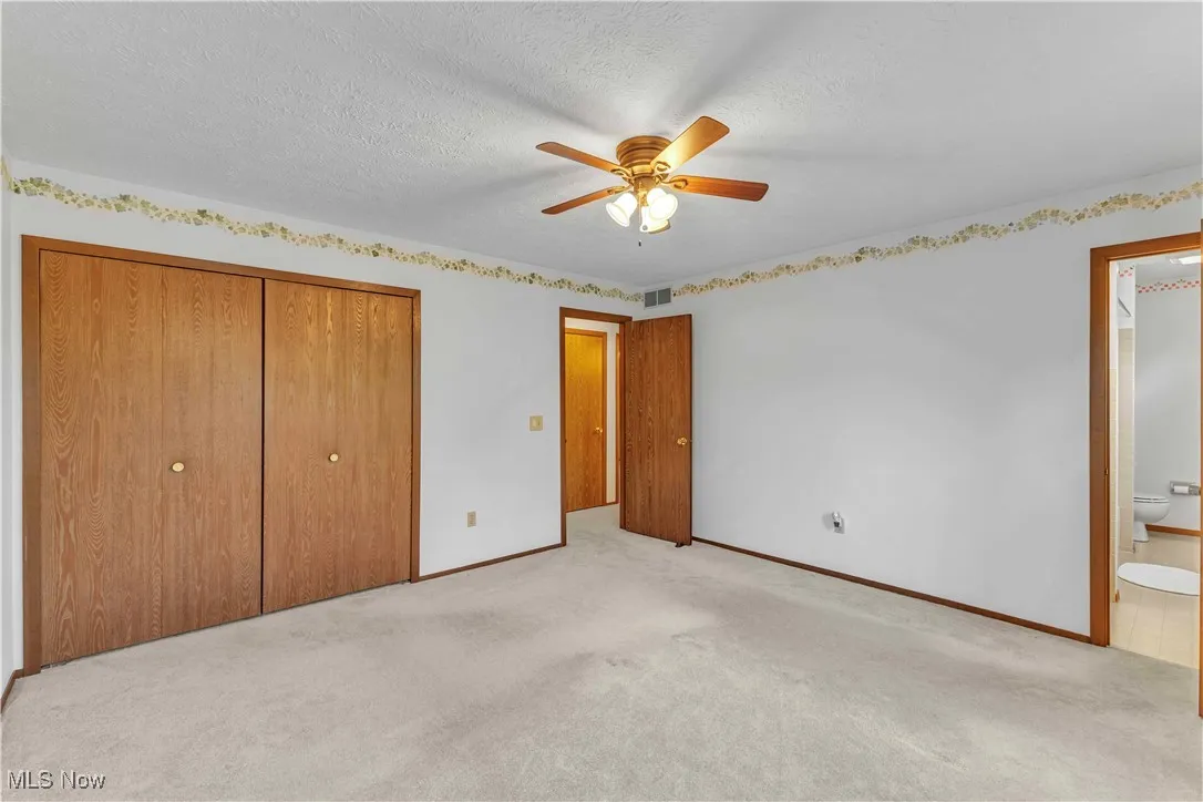 Spacious primary bedroom featuring light colored carpeting, neutral wall color, overhead ceiling fan and access to ensuite bathroom.
