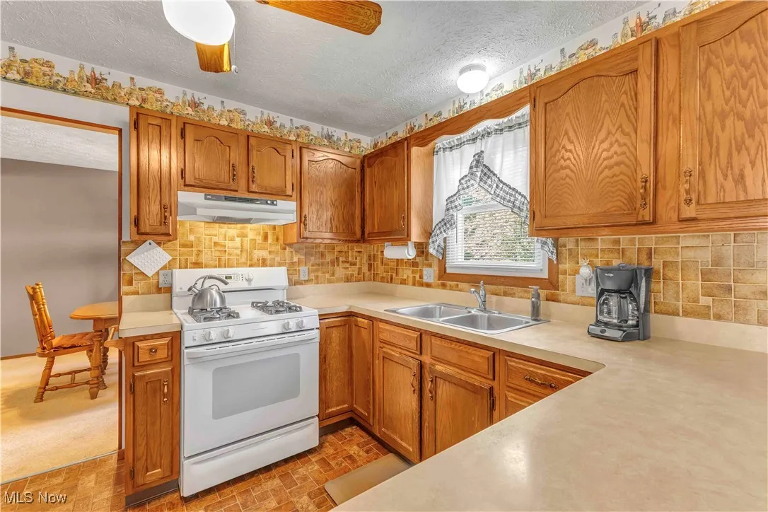 Kitchen with solid, wood cabinetry, white gas stove, white overhead range fan, kitchen sink with window above and light colored counters.