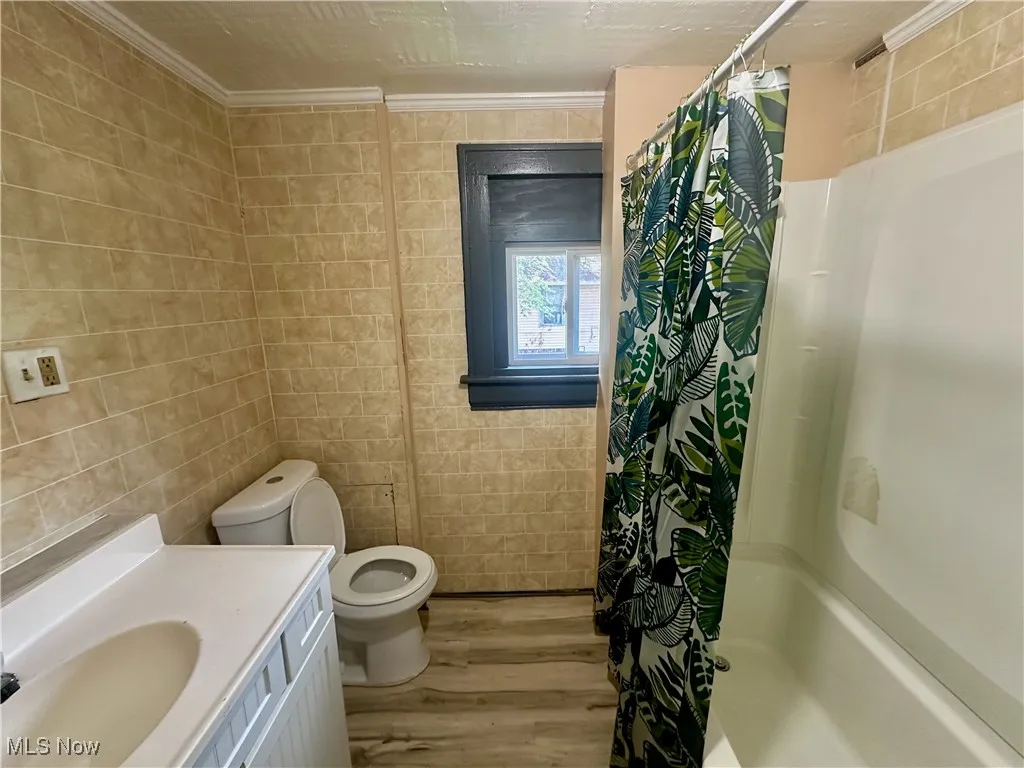 Full bath featuring tile walls, light wood-style floors, shower / bath combo with shower curtain, ornamental molding, and vanity