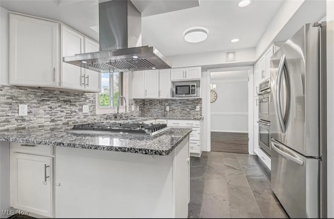 Kitchen with a peninsula, dark stone countertops, white cabinets, appliances with stainless steel finishes, and island range hood