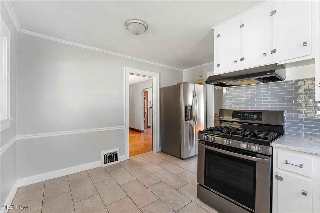 Kitchen with appliances with stainless steel finishes, white cabinets, ornamental molding, under cabinet range hood, and decorative backsplash