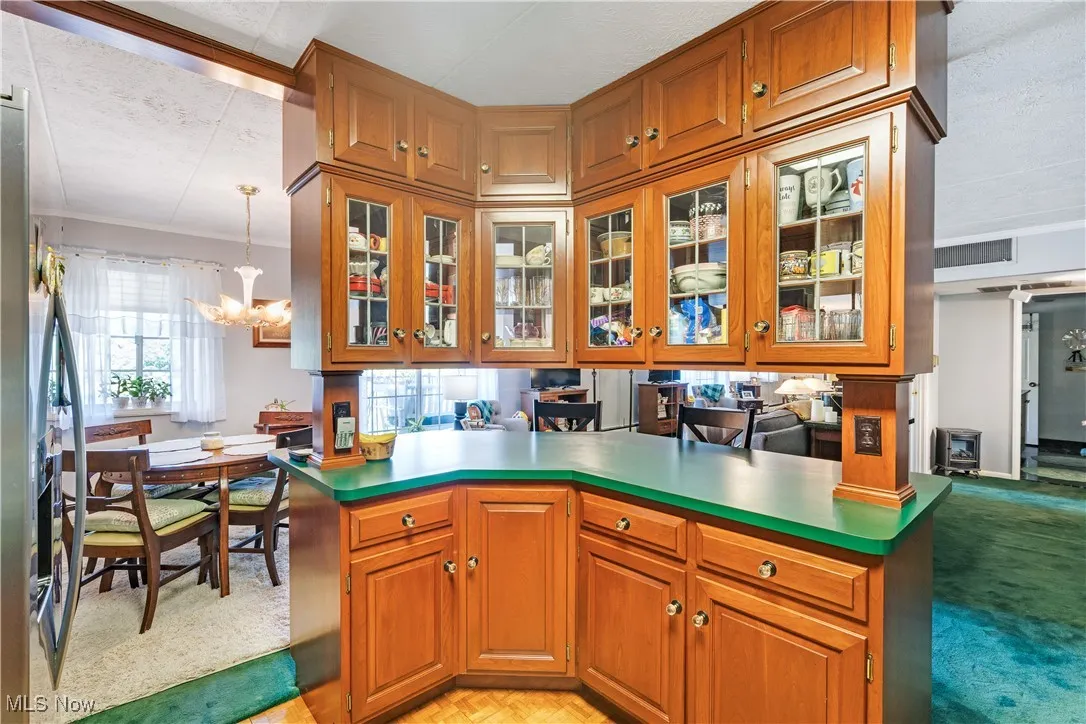 Kitchen with brown cabinetry, stainless steel refrigerator with ice dispenser, ornamental molding, hanging light fixtures, and glass insert cabinets