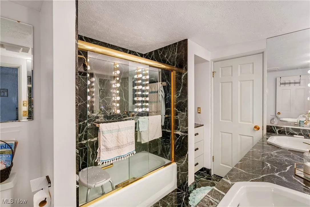 Bathroom featuring enclosed tub / shower combo, dark marble finish flooring, a textured ceiling, and double vanity