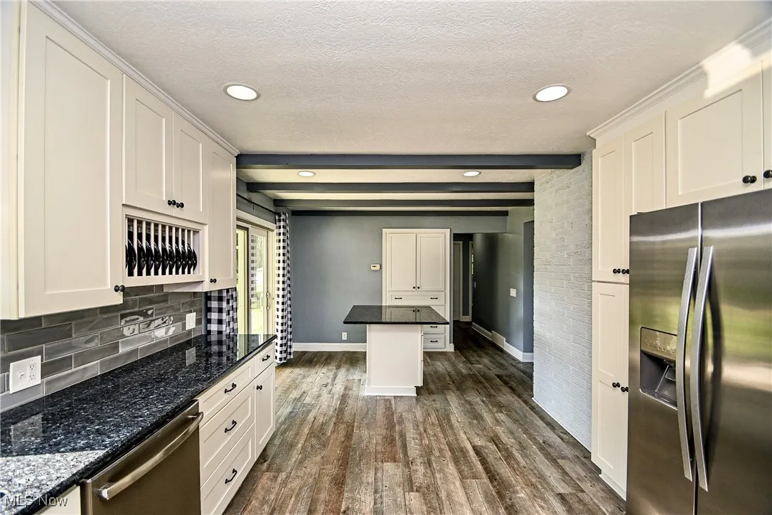 Kitchen with stainless steel appliances, beam ceiling, dark stone counters, white cabinetry, and dark wood-style flooring