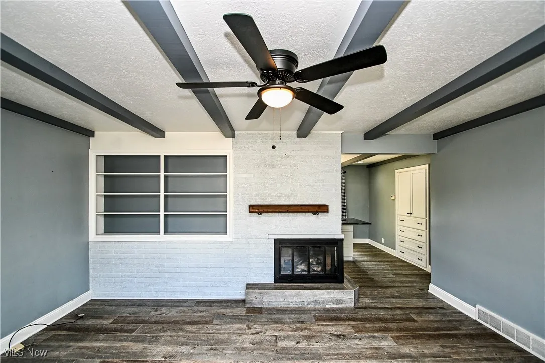 Unfurnished living room featuring a textured ceiling, beam ceiling, a glass covered fireplace, dark wood-style floors, and a ceiling fan