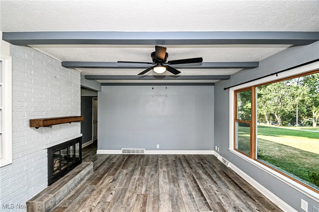 Unfurnished living room with beamed ceiling, a textured ceiling, a brick fireplace, dark wood-type flooring, and ceiling fan