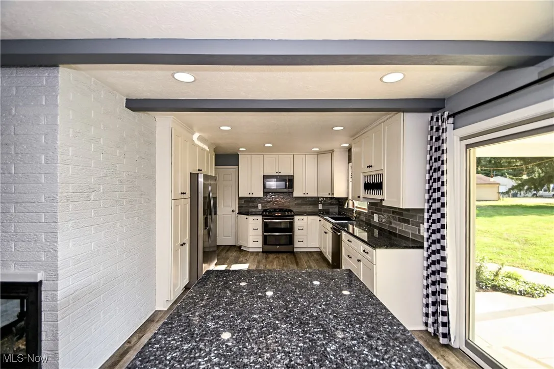 Kitchen with brick wall, stainless steel appliances, white cabinets, dark stone counters, and tasteful backsplash