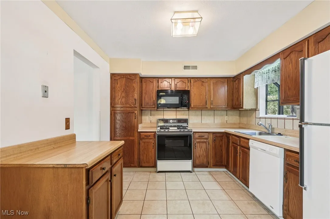 Kitchen featuring brown cabinetry, light countertops, white appliances, light tile patterned floors, and decorative backsplash