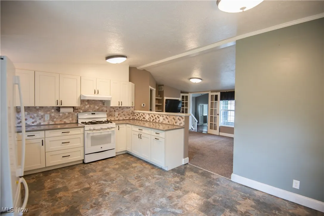 Kitchen featuring white appliances, a peninsula, tasteful backsplash, white cabinets, and dark colored carpet