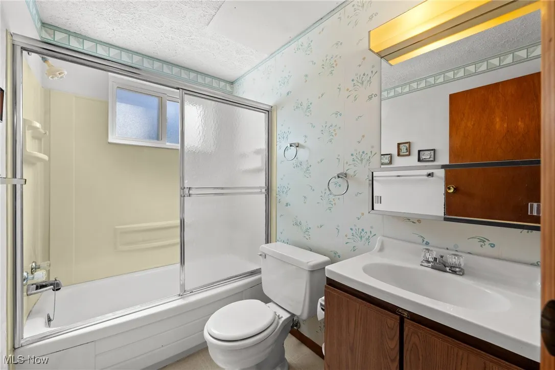Full bathroom featuring wallpapered walls, enclosed tub / shower combo, vanity, and a textured ceiling