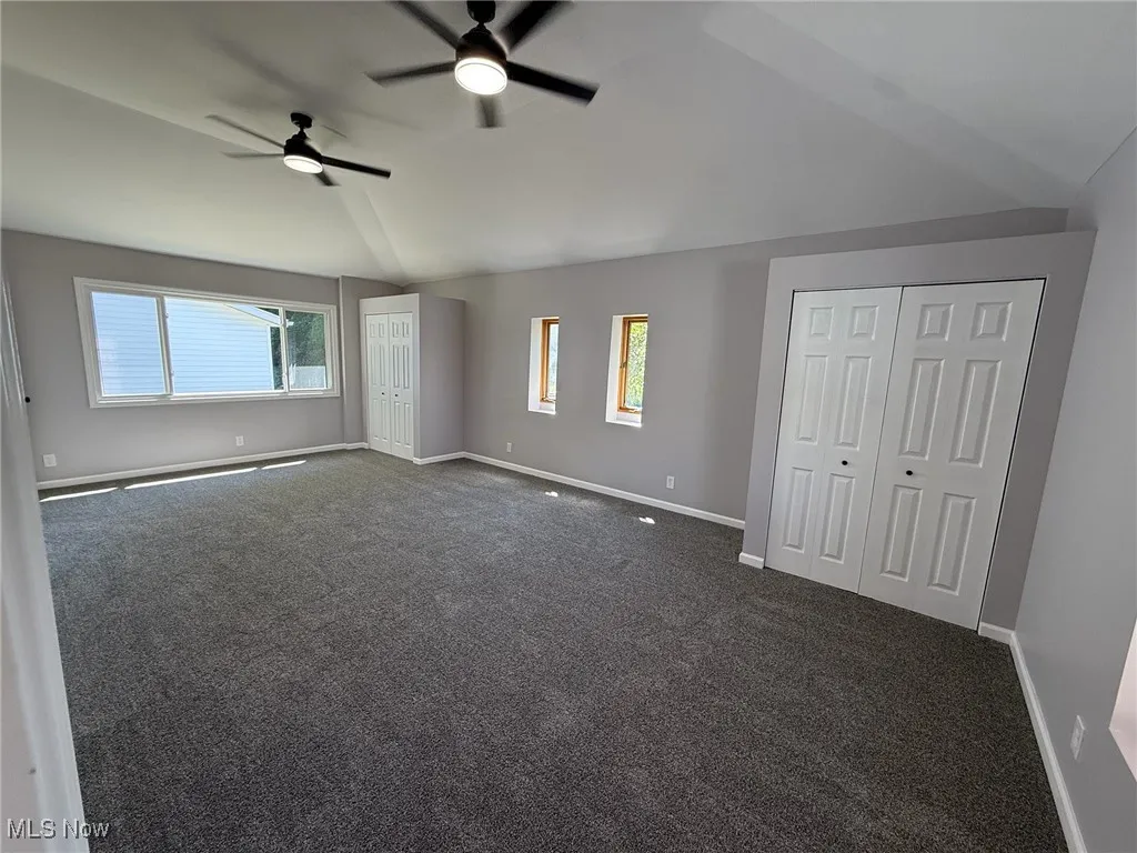 Unfurnished bedroom featuring vaulted ceiling, multiple windows, dark colored carpet, a ceiling fan, and multiple closets