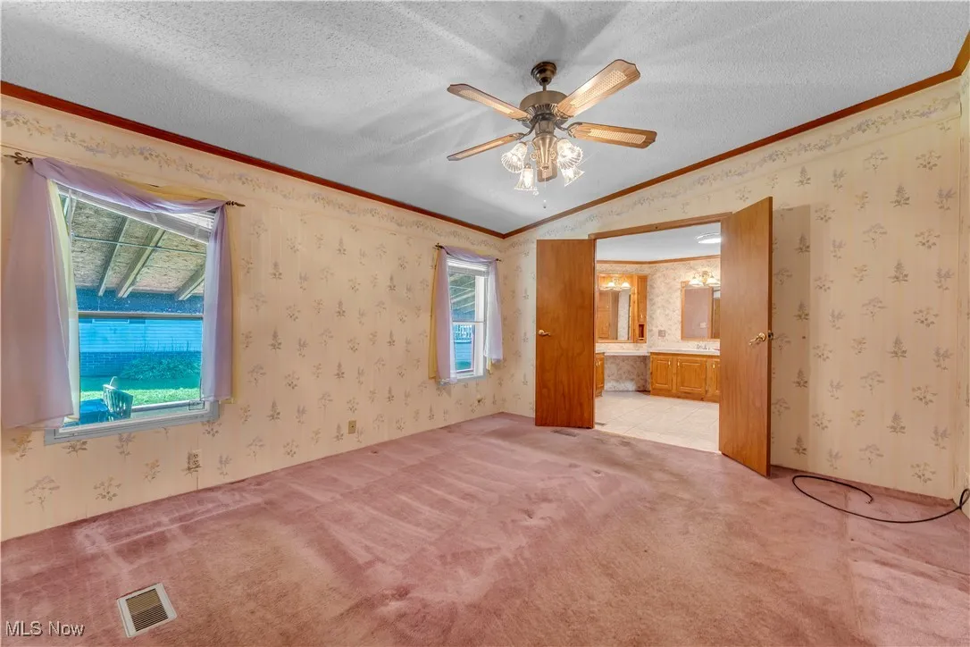 Unfurnished bedroom featuring wallpapered walls, a textured ceiling, light colored carpet, ornamental molding, and a ceiling fan